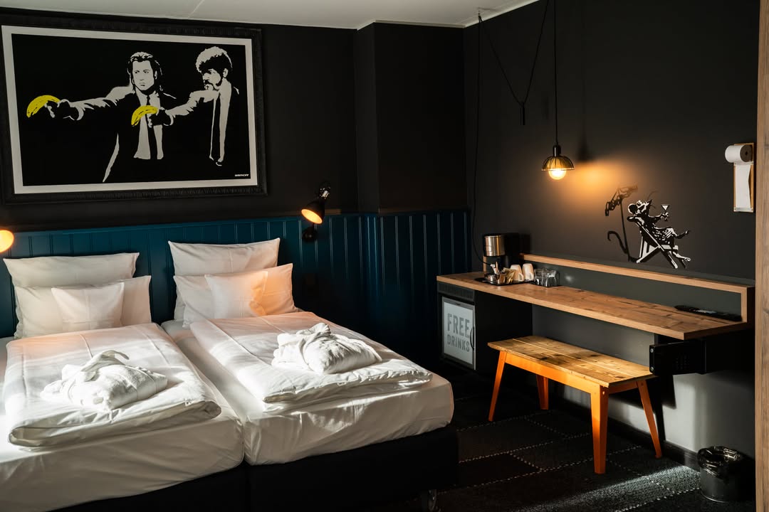 Mystery of Banksy Popup-Room Bett me and all hotel mainz. Ein gigantisches Bild hinter dem kuscheligen Boxspring-Bett zeigt das berühmte Werk „Pulp Fiction“ von 2002, eines seiner frühesten Schablonengraffitis aus London. Es zitiert den legendären Quentin Tarantino-Film, nur um den Protagonisten Vincent und Jules (John Travolta und Samuel L. Jackson) statt Waffen Bananen in die Hand zu drücken.